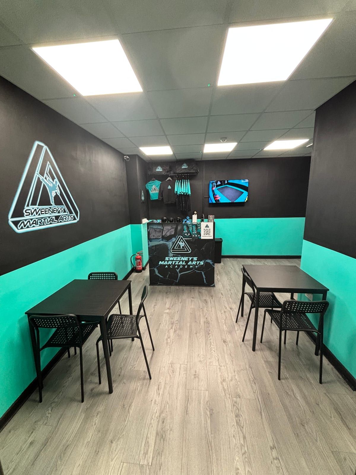 The parents’ waiting area at Sweeney’s: tables, the glowing club logo, and a screen showing the mats